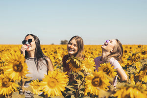 Three women laughing in a sunflower field, each with a different color of Noz reef safe facial sunscreen on their noses.